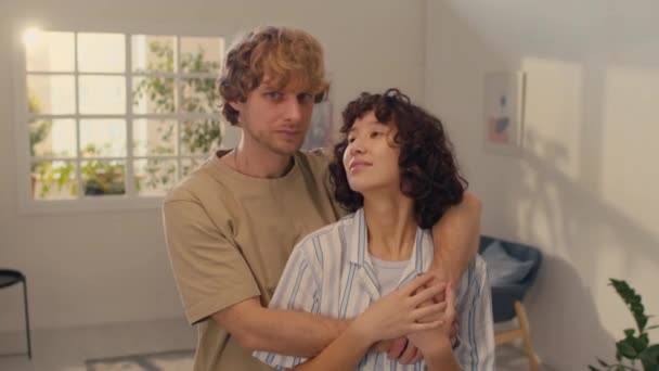 Portrait of young married couple standing in center of their living room, hugging each other while looking at camera - Footage, Video