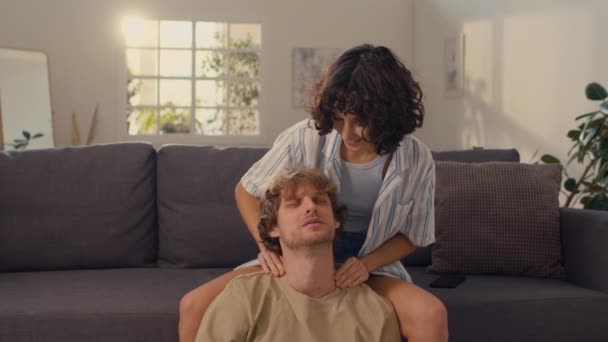 Mediana toma de mujer joven masajeando hombros de su marido mientras él sentado en el suelo relajante - Imágenes, Vídeo