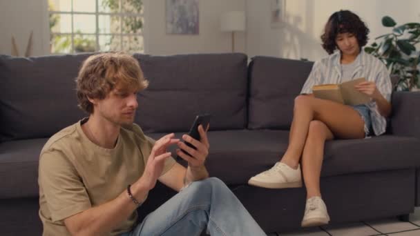Молода подружня пара, сидячи в добре освітленій вітальні разом, роблячи окремі заходи, людина пише на мобільний телефон і дівчина читає книгу на дивані - Кадри, відео