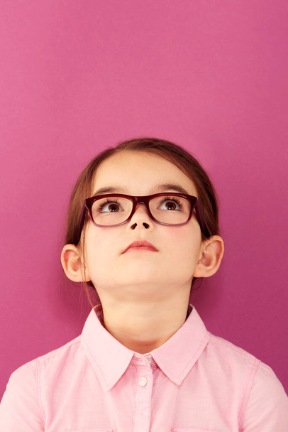 Glasses, child and looking up with thinking geek and modern fashion in a studio with serious face. Kid, eyewear and sight with outfit and nerd with accessory and pink background with confidence. - Photo, Image