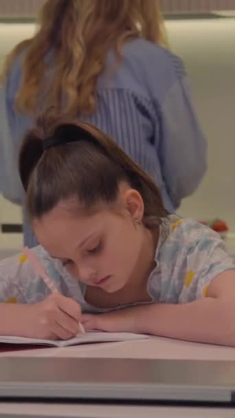Вертикальний вибірковий фокус дівчини-підлітка, що сидить на кухонному столі, робить домашнє завдання, поки її мати готує їжу на фоні - Кадри, відео