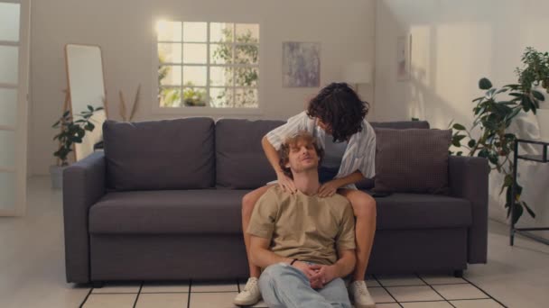 Νεαρή γυναίκα με μαύρα σγουρά μαλλιά κάθεται στον καναπέ κάνοντας μασάζ στον ώμο του φίλου της κάθεται στο πάτωμα - Πλάνα, βίντεο