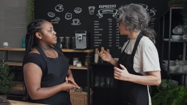 Mittlere Aufnahme einer älteren kaukasischen Coffeeshop-Mitarbeiterin, die fröhlich eine neue schwarze Barista betreut - Filmmaterial, Video