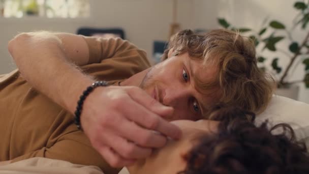 Średniej bliskości ujęcie białego młodego mężczyzny leżącego na łóżku delikatnie dotykającego twarzy swojej nierozpoznawalnej dziewczyny z czarnymi kręconymi włosami - Materiał filmowy, wideo
