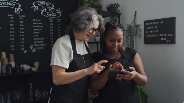 Plan moyen de deux baristas portant des tabliers noirs debout dans un café en regardant des photos sur le téléphone - Séquence, vidéo