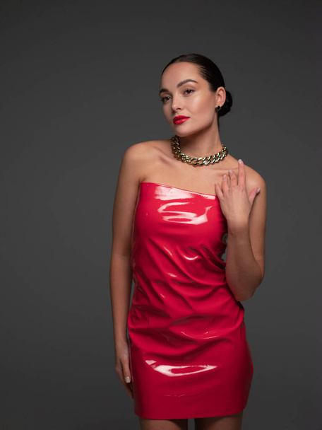 Young pretty brunette woman poses in stylish red latex dress and chain in her neck in studio against gray background. - Photo, Image