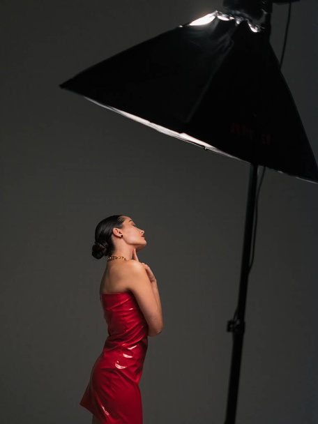 Молода красива брюнетка позує в стильній червоній латексній сукні під прожектором в студії на сірому фоні. - Фото, зображення