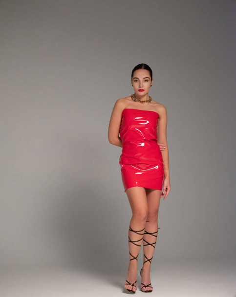 Young pretty brunette woman poses in stylish red latex dress and high heels sandals with straps in studio against gray background. - Фото, изображение