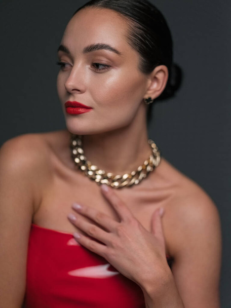 Portrait of young beautiful brunette woman in red latex dress with chain in her neck and red lipstick. Cosmetology, cosmetics, skin care, natural beauty. - Photo, Image