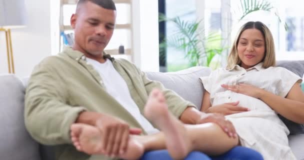 O homem esfrega os pés de sua esposa grávida enquanto relaxa em um sofá na sala de estar de sua casa familiar. Calma, descanso e jovem mulher com gravidez deitada no sofá com uma pessoa masculina tocando suas pernas - Filmagem, Vídeo