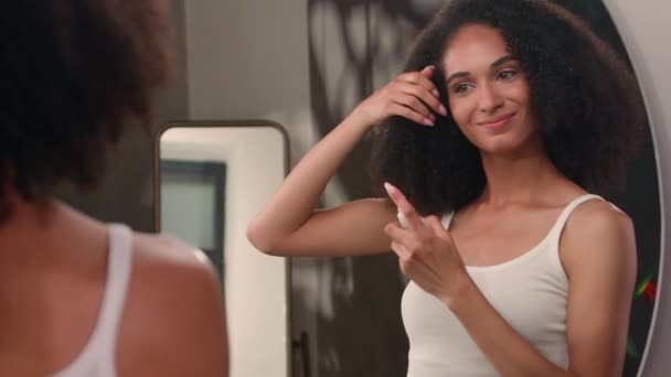 donna afro-americana guardando specchio riflesso ragazza femminile in bagno di casa applicare lacca per capelli cosmetici liquidi per la cura dei capelli twist ricci acconciatura cura acconciatura bellezza routine - Filmati, video
