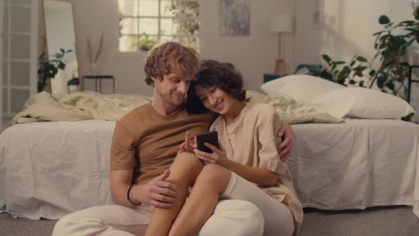 Πλήρης εικόνα του παντρεμένου ζευγαριού κάθεται άνετα στο πάτωμα του υπνοδωματίου τους δίπλα στο κρεβάτι τους κοιτάζοντας το τηλέφωνο στα χέρια της γυναίκας με μαύρα μαλλιά - Πλάνα, βίντεο