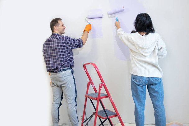 ローラー ブッシュを握るカップルはバイオレット色のペンキ新しい家のアパートのリビング ルームの壁を塗ります. - 写真・画像