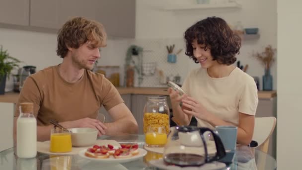 シリアルとサンドイッチで作られた朝食を持ってキッチンに座っている若いカップル - 映像、動画