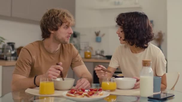 Średnie ujęcie młodej pary rozmawiającej ze sobą przy stole jedzącej płatki z mlekiem i sokiem pomarańczowym - Materiał filmowy, wideo
