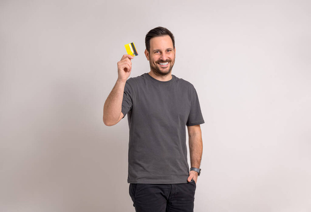 Retrato de joven vendedor sonriente con la mano en el bolsillo que muestra la tarjeta de crédito sobre fondo blanco - Foto, Imagen