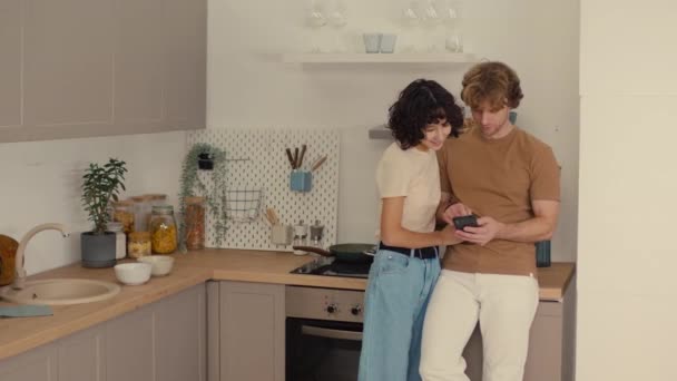 Joven de pie en la cocina mirando a través de las redes sociales en su teléfono y compartiéndolo con su novia - Metraje, vídeo