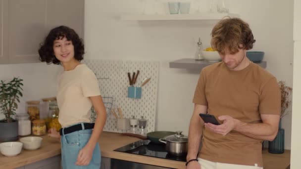 Średnie ujęcie młodej kobiety z ciemnymi kręconymi włosami sprzątającej w kuchni, podczas gdy jej chłopak przewija się przez telefon - Materiał filmowy, wideo