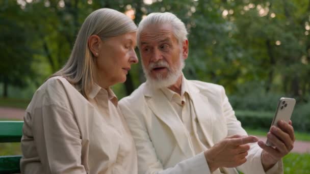 Elderly Caucasian gray husband man hold showing mobile phone explaining discuss talking aged wife woman outside city park. Grandparents pensioners old family learning modern online technologies gadget - Footage, Video