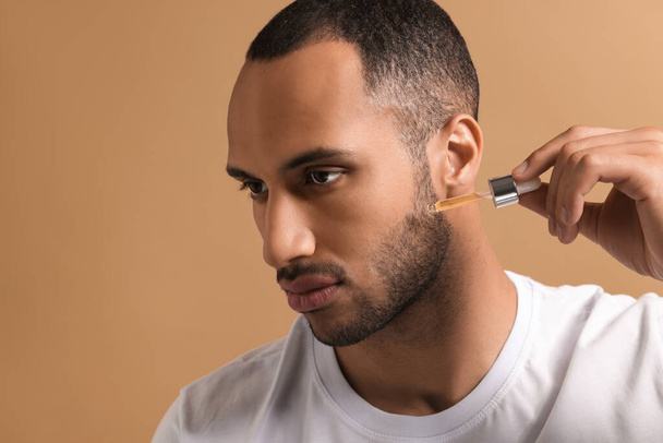 Handsome man applying cosmetic serum onto face on light brown background, closeup. Space for text - Foto, Imagem
