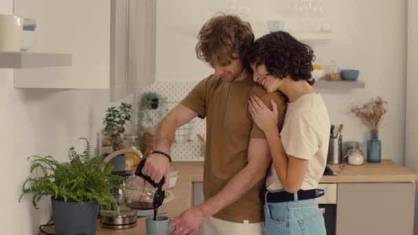 Νεαρό ζευγάρι στέκεται στην κουζίνα αγκαλιάζοντας ο ένας τον άλλον, ενώ ο άνθρωπος ρίχνει καφέ στην κούπα του - Πλάνα, βίντεο