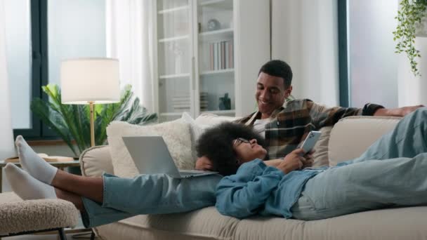 Evdeki kanepede rahat bir şekilde oturan Afrikalı Amerikalı çift dizüstü bilgisayarlı kadını kullanan, internetteki alet bağımlısı adam internette sosyal medya bağımlılığı sitelerinde cep telefonlarını karıştırıyor. - Video, Çekim