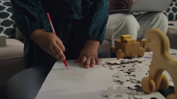 Midsection toma de niño irreconocible en vestido verde dibujo de dibujos con lápiz rojo mientras su padre sentado en el sofá detrás de ella - Metraje, vídeo