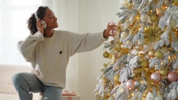 Vrolijk kerstfeest. Afro-Amerikaanse vrouw met koptelefoon die muziek afluistert die kerstboom versiert. Gelukkig meisje bij klassieke traditionele kerstboom. Kerstavond thuis tijd voor feest - Video