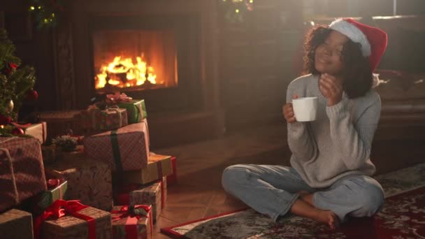 Feliz Navidad. Mujer afroamericana en sombrero de Santa con taza de té de café de bebida caliente cerca del árbol de Navidad en el interior oscuro. Chica en la sala de estar con árbol de Navidad y chimenea. Nochebuena en casa - Metraje, vídeo