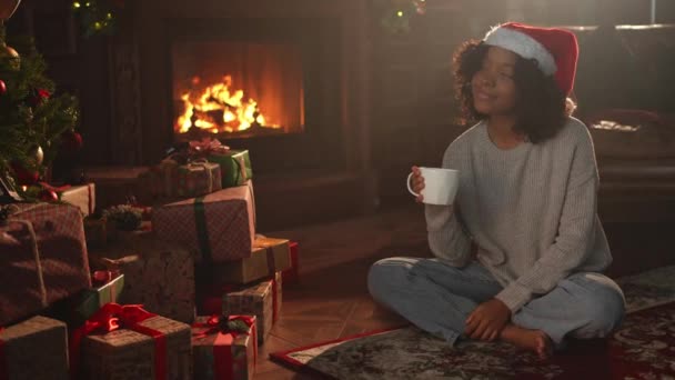 Veselé Vánoce. Africká Američanka v klobouku Santa s šálkem horkého nápoje káva čaj u vánočního stromku v tmavém interiéru. Dívka v obývacím pokoji s vánočním stromečkem a krbem. Štědrý večer doma - Záběry, video