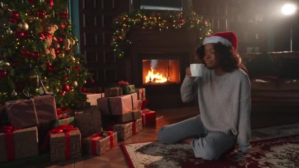 メリークリスマス。 サンタハットのアフリカ系アメリカ人女性は,暗いインテリアでクリスマスツリーの近くでホットドリンクコーヒーティーを飲みます. クリスマスツリーと暖炉のあるリビングルームの女の子. 自宅でクリスマスイブ - 映像、動画