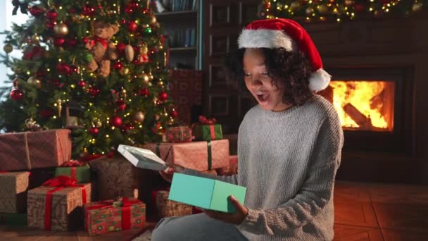 Boldog karácsonyt! Az afro-amerikai nő kipakolja az ajándékdobozt a karácsonyfa közelében. Lány a nappaliban karácsonyfa és kandalló nyitó ajándék doboz meglepetés arcát. Karácsony este otthon. - Felvétel, videó