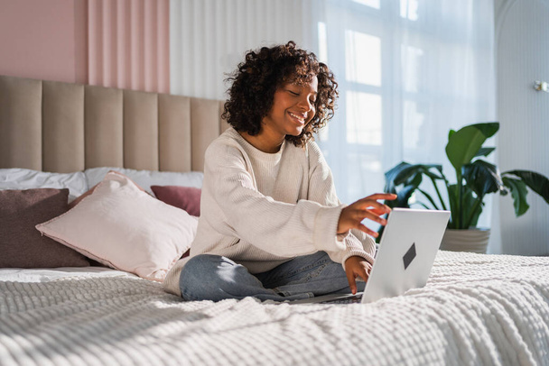 Afroamerykanka używająca laptopa w łóżku w biurze, pisząca na czacie, czytająca maile. Młoda czarna kobieta ma wirtualne spotkanie online czat wideo telekonferencja. Kształcenie zawodowe w domu - Zdjęcie, obraz