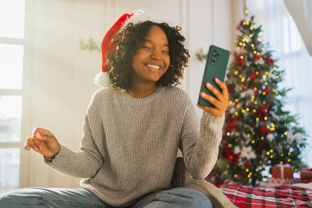 Feliz Navidad. Mujer africana en Santa sombrero de tener chat de vídeo en el teléfono inteligente. Blogger en el humor festivo grabación de vídeo en la red social. Chica teniendo videollamada con amigos familia celebrando la Navidad - Foto, Imagen