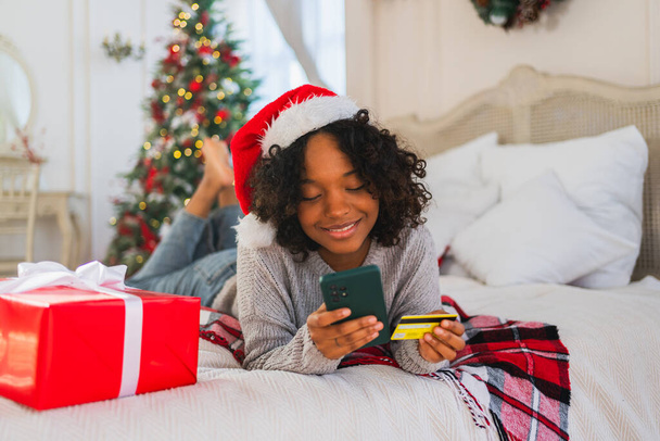 African American woman shopping online holding smartphone paying with gold credit card for Christmas gifts. Girl buying on Internet enter credit card details enjoying Christmas winter holidays at home - Photo, Image