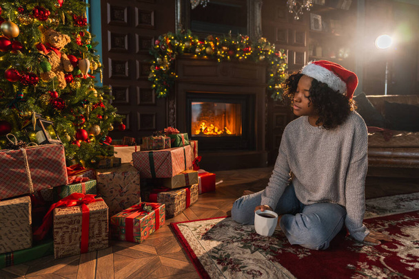 Merry Christmas. African American woman in Santa hat with cup of hot drink coffee tea near Christmas tree in dark interior. Girl in living room with Christmas tree and fireplace. Christmas eve at home - Photo, Image
