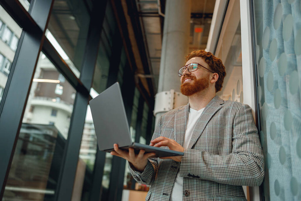 Glimlachende mannelijke sales manager in bril werkend op laptop staande in het kantoor tijdens de werkdag - Foto, afbeelding