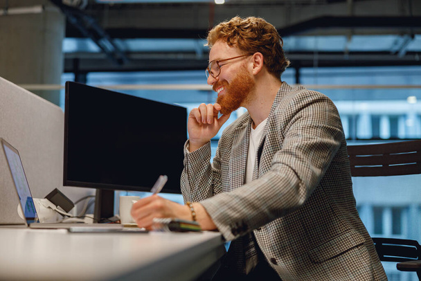 Χαμογελώντας αρσενικό διαχειριστή που εργάζονται σε φορητό υπολογιστή, ενώ κάθεται στο γραφείο στο φόντο του γραφείου  - Φωτογραφία, εικόνα