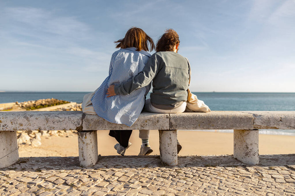 Вид сзади на пару объятий, сидя на скамейке и наслаждаясь прекрасным видом на океан. Высокое качество фото - Фото, изображение