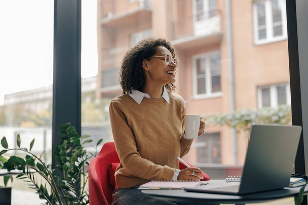 Hymyilevä nainen yrittäjä työskentelee kannettavan tietokoneen, muistiinpanoja kodikas coworking tilaa sisustus - Valokuva, kuva