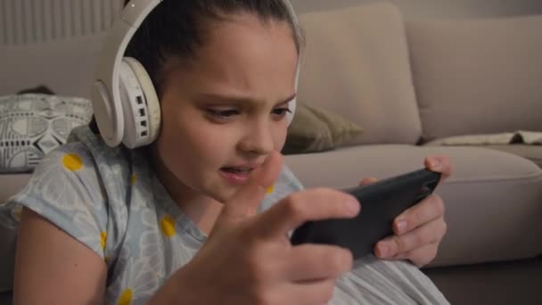 Ψηφιακή μητρική παιδί με ασύρματα ακουστικά πάρει αναστατωμένος μετά την απώλεια στο κινητό παιχνίδι στο gadget της - Πλάνα, βίντεο