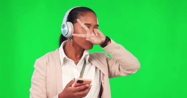 Business woman, phone and headphones on green screen for music, dancing and peace sign. Female model person with smartphone for listening to music with fun energy, smile and motivation in studio. - Footage, Video