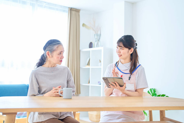 Νεαρή γυναίκα με ποδιά και ηλικιωμένη γυναίκα συζητούν στο σπίτι - Φωτογραφία, εικόνα
