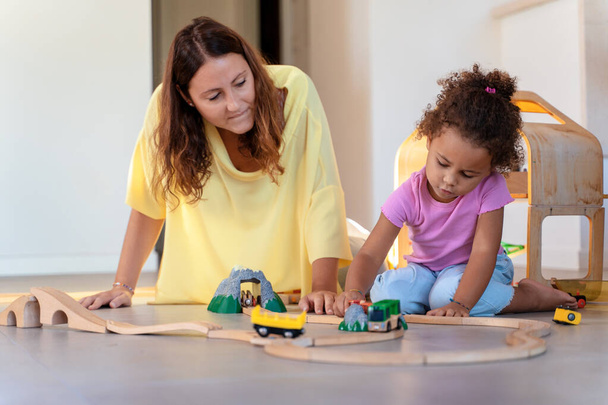 Madre e hija interraciales se centran en la construcción de una vía de tren de madera, disfrutando de juegos creativos juntos en interiores. - Foto, imagen