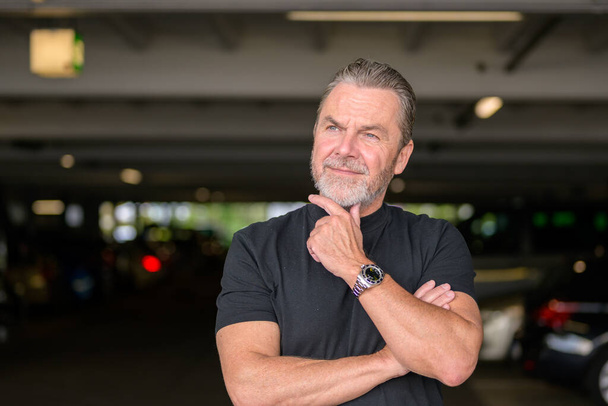 Привабливий сіро-волохатий чоловік з чорною футболкою носить стильний годинник і дивиться в бік автостоянки - Фото, зображення