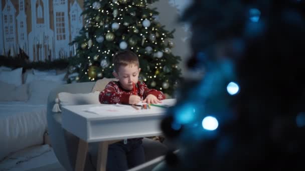 Karácsonyi kívánságok: Kisfiú levelet komponál a Mikulás az asztalnál, boldog várakozást ünnepélyesen díszített otthon. Kiváló minőségű 4k felvételek - Felvétel, videó