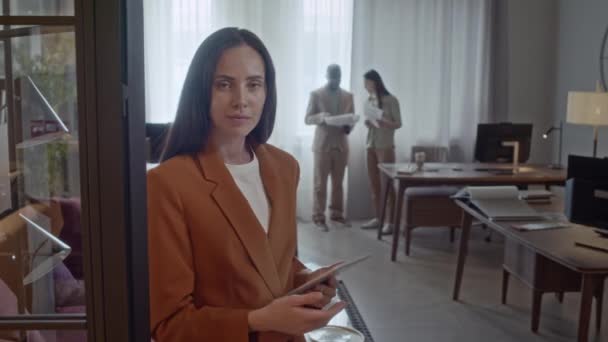 Portrait of Caucasian female corporate worker leaning against doors of companys office, holding digital tablet in her arms, looking at camera - Footage, Video