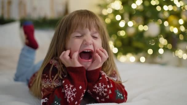 Portret van een lachend blank meisje op een bed in de buurt van een kerstboom. Gelukkig kind op nieuwjaarsvakantie thuis. Hoge kwaliteit 4k beeldmateriaal - Video