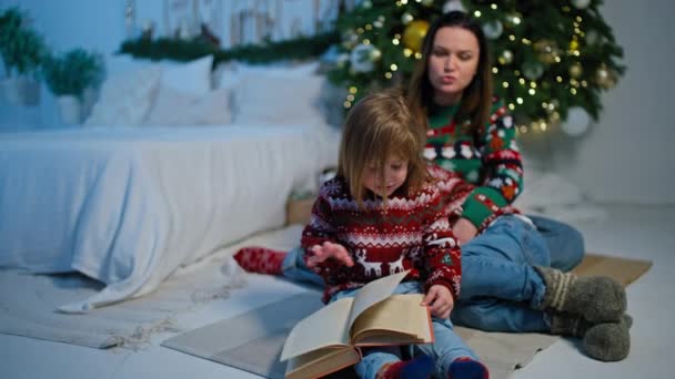 Varázslatos mesék: Anya és a gyerekek karácsonyi történeteket olvasnak a fa mellett, ünnepi hangulatot teremtve ünnepi dekorációkkal otthon. Kiváló minőségű 4k felvételek - Felvétel, videó