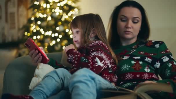 Örömteli ünnepségek: Anya és lánya közös nevetés, néz karácsonyi rajzfilmek telefonon, átöleli az újévi szellem otthon. Kiváló minőségű 4k felvételek - Felvétel, videó